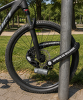 Candado U-Lock Odis K1800D gris de alta seguridad y cadena antirrobo de eslabones gruesos con funda textil negra fijando la rueda delantera de una bicicleta a un poste urbano. Fotografía de uso en exterior distribuida por Proyecto Madera Chile.
